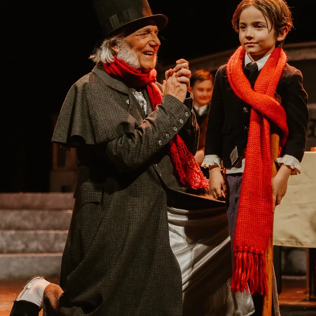 Two actors on stage. Old man in top hat and red scarf and young boy in red scarf depicting Scrooge and Tiny Tim from "A Christmas Carol."