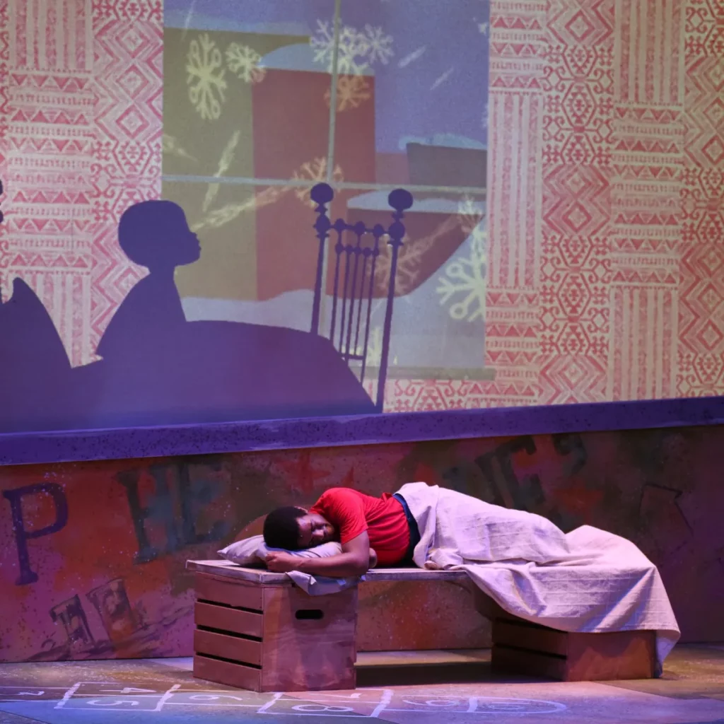 Person dressed in red sleeps in a bed on a stage with an image of a boy awake in bed projected on the stage behind them.