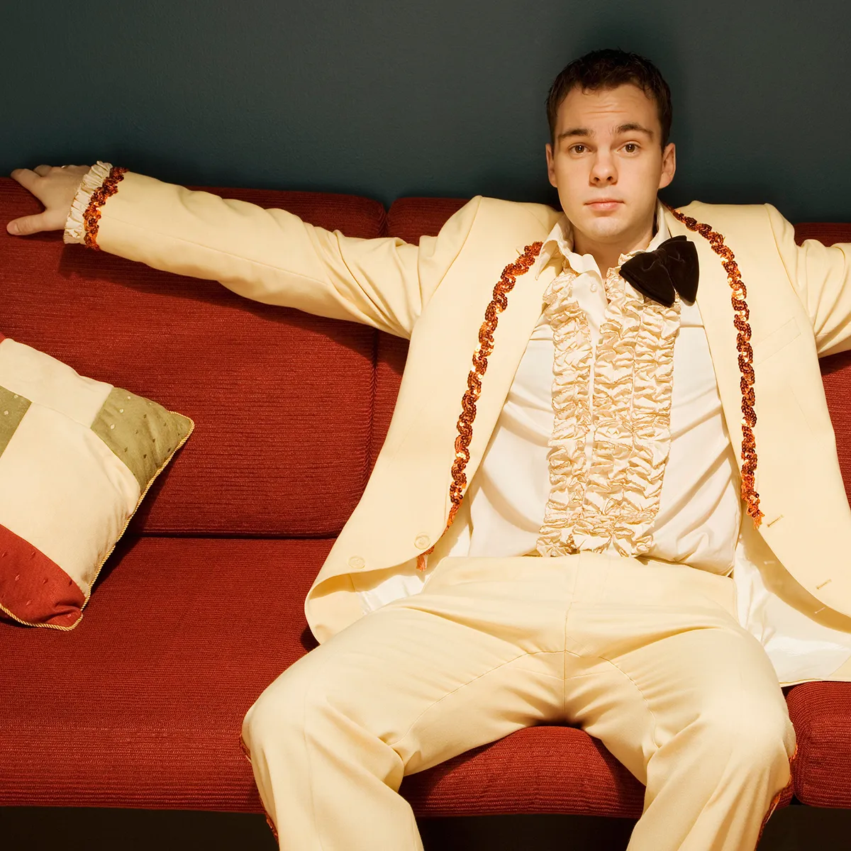 Young man wearing a cream-colored , ruffled vintage tuxedo with the bowtie undone stretches his arm out on the back of the couch he is sitting on.