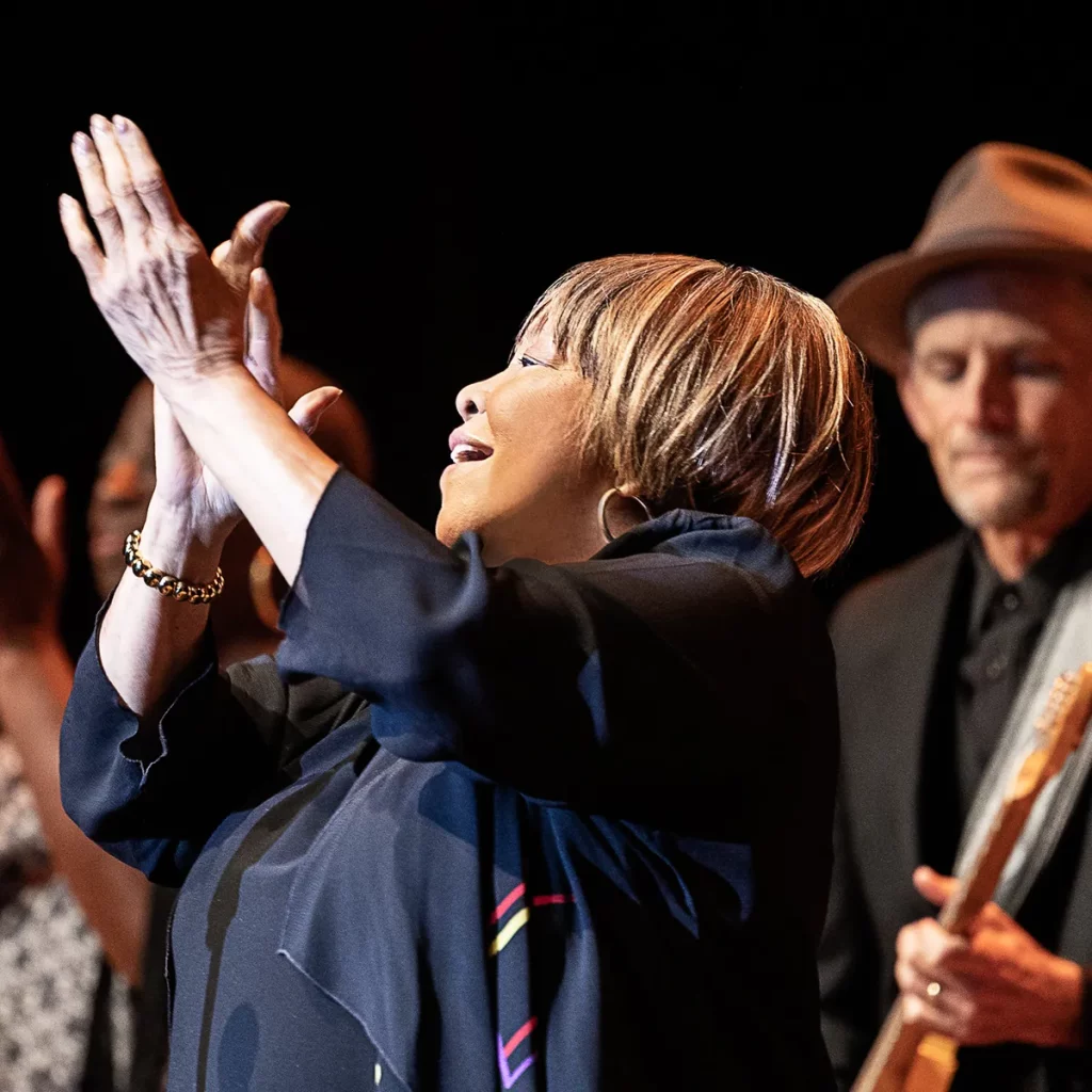 Photo of Mavis Staples on stage with band clapping her hands.