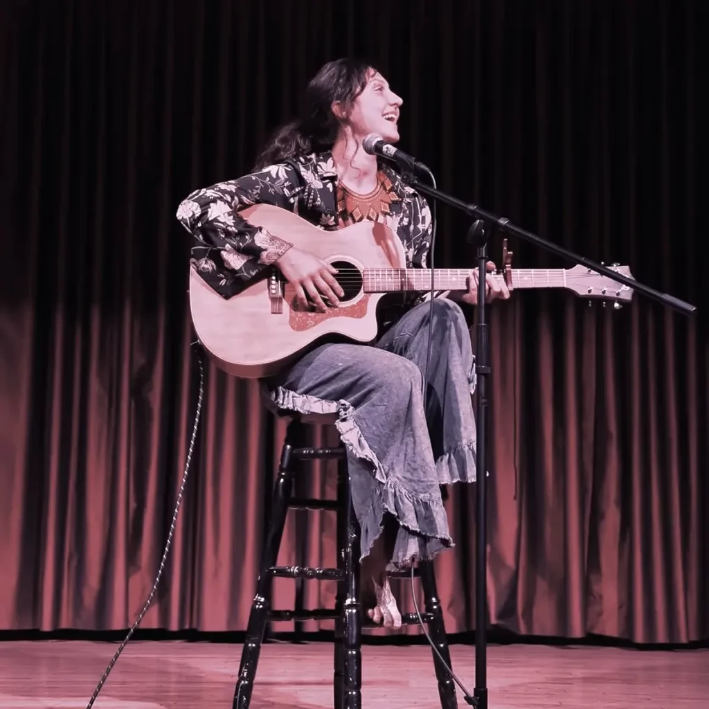 Musician Lydia Cooley, sitting on a stool on stage with guitar.