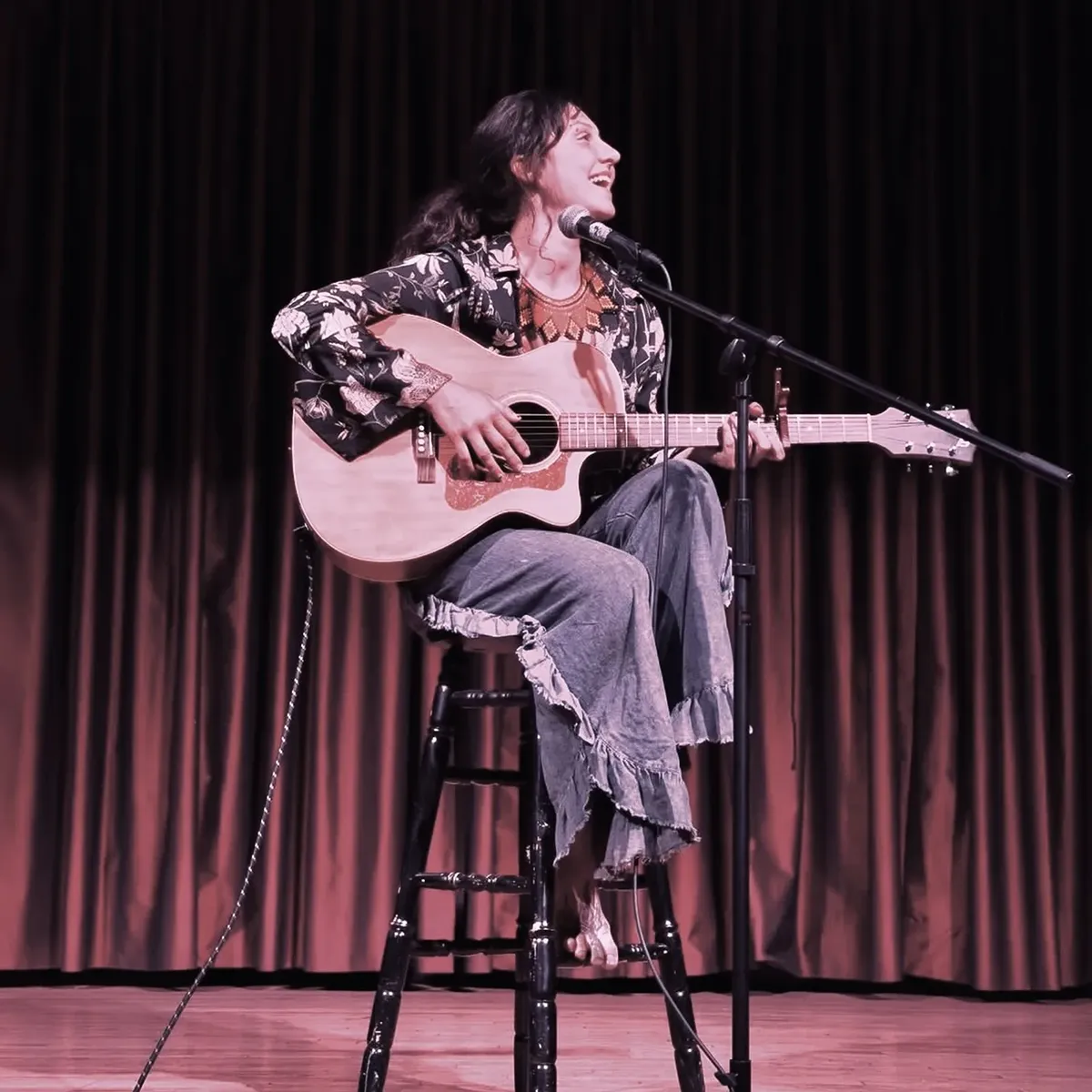 Musician Lydia Cooley, sitting on a stool on stage with guitar.